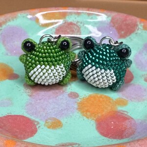 May include: Two beaded frog keychains. One is light green, the other is dark green. Both have white bellies and black eyes. The keychains are on a silver ring. The frogs are sitting on a speckled, light green ceramic plate.