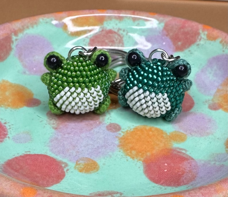 May include: Two beaded frog keychains. One is light green, the other is dark green. Both have white bellies and black eyes. The keychains are on a silver ring. The frogs are sitting on a speckled, light green ceramic plate.