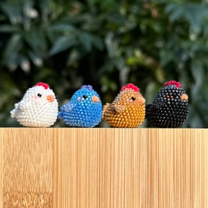 May include: Four small beaded chicken figurines, each with a different colour: white, blue, brown, and black. The chickens are lined up on a wooden surface.