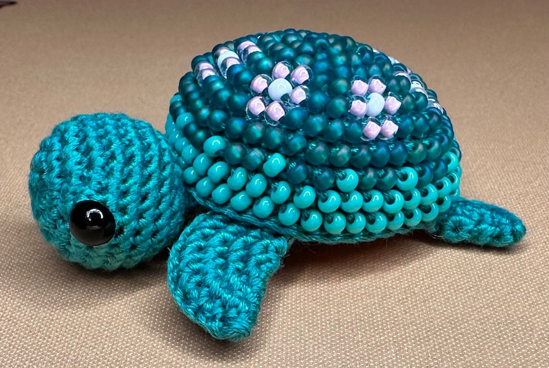 May include: A handcrafted turquoise crochet turtle with a beaded shell. The turtle's shell is made of teal, blue, and white beads. The turtle has a black eye and is a unique decorative item.