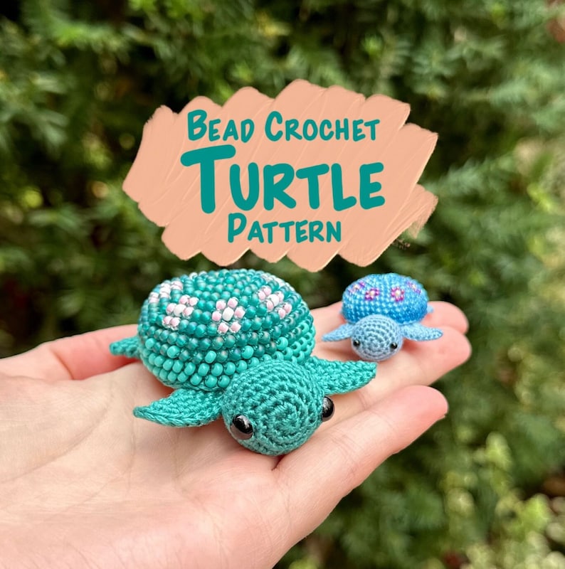 May include: A hand holds two crocheted turtle figurines. The larger turtle is teal with white and pink floral accents. The smaller turtle is light blue with a blue and pink floral pattern. The image includes the text "Bead Crochet Turtle Pattern."