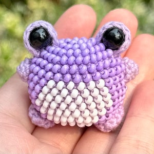 May include: A handmade, beaded frog figurine in shades of lavender and white. The frog has large, black eyes and is crafted with small beads. The frog's body is a mix of lavender and white beads, creating a unique texture and design.