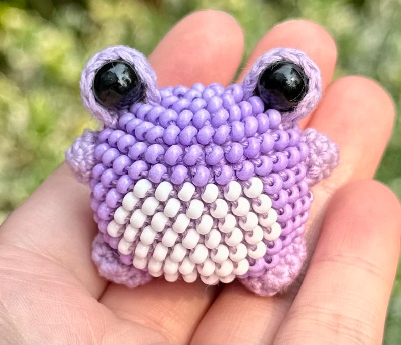 May include: A handmade, beaded frog figurine in shades of lavender and white. The frog has large, black eyes and is crafted with small beads. The frog's body is a mix of lavender and white beads, creating a unique texture and design.