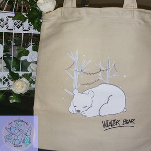 May include: A beige canvas tote bag featuring a white polar bear sleeping in a snowy forest scene. The text "WINTER BEAR" is written below the bear.