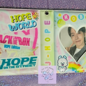 Peut inclure: Un porte-cartes photo en plastique transparent avec une découpe en forme de cœur. Le support présente du texte et des graphiques colorés, dont "HOPE WORLD", "JACK IN THE BOX" et "HOPE ON THE STREET". Une photo d'une personne est visible.
