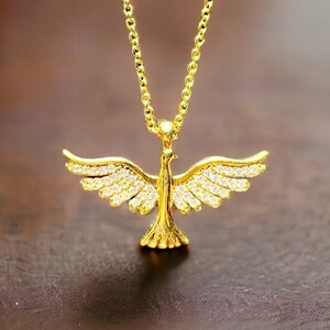 May include: A gold-colored necklace featuring a dove pendant with outstretched wings. The dove is adorned with small, sparkling stones. The chain and pendant are displayed against a dark, blurred background, highlighting the jewelry's details.