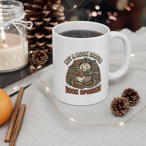 May include: A white ceramic mug featuring a colorful illustration of an opossum sitting on a large stack of books. The text above and below the opossum reads, "NOT A BOOK DRAGON" and "BOOK OPOSSUM" in a brown, rounded, retro-style font.
