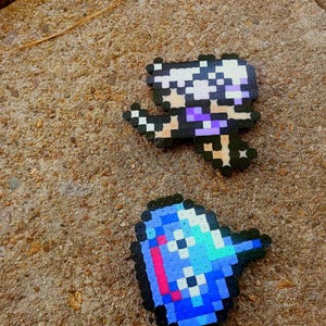 May include: Two pixel art figures made from colorful plastic beads. One figure is a person with white hair and purple clothing. The other is a blue and pink pixelated object with a black outline. Both figures are on a textured, brown surface.