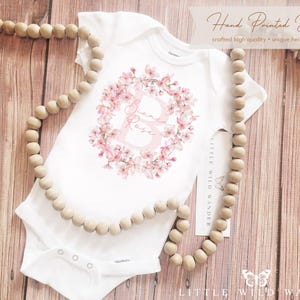 May include: White baby onesie with a floral wreath design in pink, featuring the name "Biane Rose" in a soft pink font. The onesie is displayed on a wooden surface with a beaded garland. Text reads "Hand Printed Designs".