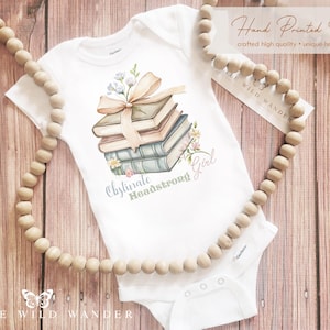 May include: White baby onesie with a watercolor illustration of a stack of books tied with a ribbon and flowers. The text "Obstinate Headstrong Girl" is printed below the books. A wooden bead garland frames the onesie.