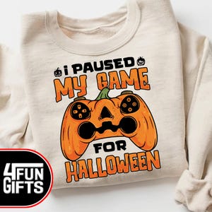 May include: A cream-colored sweatshirt with the text "I paused my game for Halloween" and a pumpkin-shaped video game controller graphic.