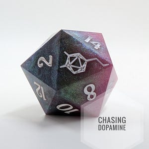 Puede incluir: Un dado D20 de resina iridiscente con numeración blanca y un diseño de molécula de dopamina. El dado presenta un degradado de color azul, verde y rosa. El texto "CHASING DOPAMINE" es visible en un hexágono blanco.