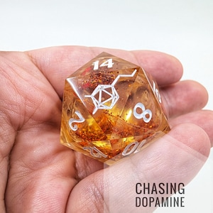 Puede incluir: Un dado transparente naranja de 20 caras con numeración blanca y un diseño de molécula de dopamina. El dado está lleno de purpurina naranja y se sostiene en una mano. El texto "CHASING DOPAMINE" es visible.