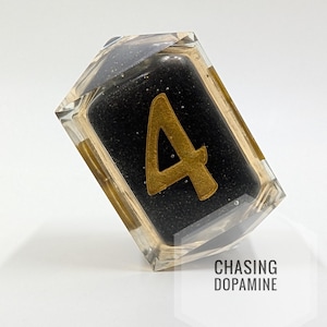 Puede incluir: Un bloque de resina transparente y rectangular con un interior negro y el número 4 dorado. El bloque tiene detalles dorados y el texto "CHASING DOPAMINE". El objeto está inclinado sobre un fondo blanco.
