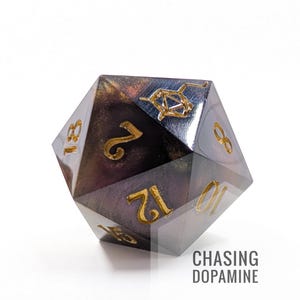 Puede incluir: Un dado de múltiples caras con números dorados y un diseño oscuro y arremolinado. El texto "CHASING DOPAMINE" está impreso en la parte inferior derecha. Este dado inusual es probablemente para juegos de mesa.