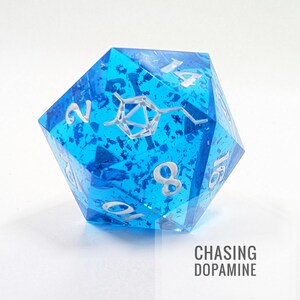 Puede incluir: Dado azul de múltiples caras con numeración plateada y un diseño de molécula de dopamina plateada. El dado tiene una apariencia texturizada y moteada. El texto "CHASING DOPAMINE" está impreso en negro en la parte inferior derecha.