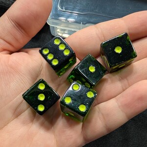 Può includere: Sono presentati cinque dadi neri con punti giallo brillante. I dadi sono trasparenti, con una tonalità verde scuro. I dadi sono in varie posizioni, mostrando diversi numeri di punti. I dadi sono probabilmente usati per i giochi.