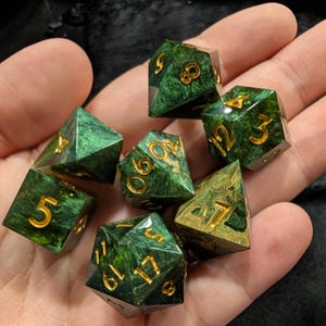 Puede incluir: Un conjunto de siete dados poliédricos verdes con números dorados. Los dados se muestran en una mano. Son para juegos de mesa. El fondo es oscuro. Los dados tienen diferentes formas, incluyendo un d20 y un d6.