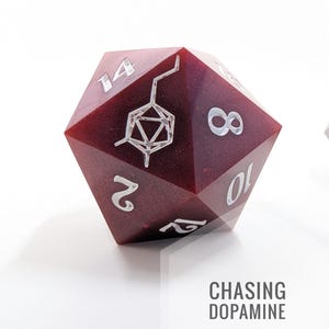 Puede incluir: Un dado D20 burdeos con numeración plateada y un gráfico plateado. El dado tiene el texto "CHASING DOPAMINE" en fuente gris.