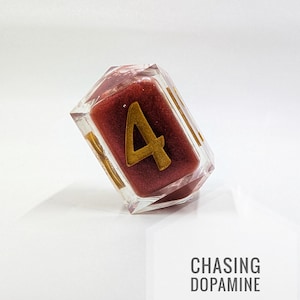 Puede incluir: Un dado transparente y multifacético con un interior burdeos y detalles dorados. El número "4" se muestra prominentemente en dorado. El texto "CHASING DOPAMINE" es visible en un hexágono blanco.