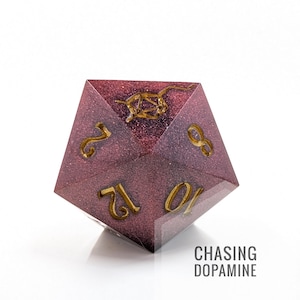 Puede incluir: Un dado D20 burdeos y dorado con numeración dorada y un emblema dorado en la parte superior. El dado tiene un aspecto brillante y moteado. El texto "CHASING DOPAMINE" está impreso en blanco en la parte inferior derecha.