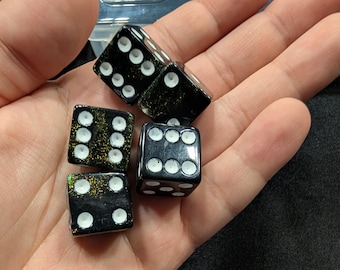 Pode incluir: Seis dados pretos com pontos brancos, segurados em uma mão. Os dados têm um efeito de brilho dourado. Estes dados são usados para jogos de tabuleiro e jogos de azar. Cada dado mede aproximadamente 1,6 cm.