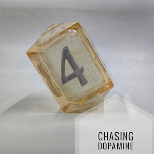 Puede incluir: Un bloque de resina rectangular transparente con un número 4 gris incrustado. El bloque tiene un tono ámbar claro y está en ángulo. El texto "CHASING DOPAMINE" se muestra en un hexágono blanco.