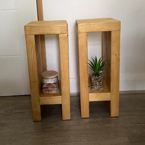 May include: Two wooden side tables with a natural finish. Each table has a rectangular top and a lower shelf. One table holds a candle in a glass jar, and the other holds a small potted plant. The tables are approximately 30 cm wide and 30 cm deep.