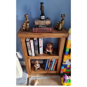 May include: A wooden bookshelf with two shelves, displaying books and decorative items. The top shelf holds cat figurines, books labelled "Curses," "Spells," and "Potions," and an owl figurine. The lower shelf contains books and bear figurines.