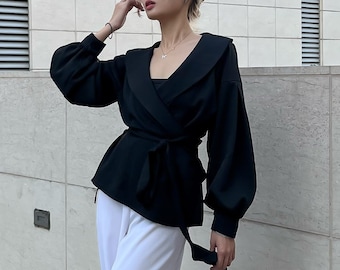 Puede incluir: Una chaqueta negra de vestir con cuello ancho y mangas largas. La chaqueta tiene un lazo en la cintura y se lleva sobre una blusa blanca.