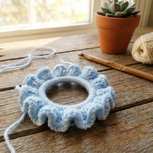 Op de afbeelding: Een lichtblauwe, handgemaakte gehaakte haarband met een gerimpelde rand. Het haaraccessoire is cirkelvormig, met een zichtbare elastische band in het midden. De achtergrond omvat een houten haaknaald, een bol wol en een vetplant in een pot.