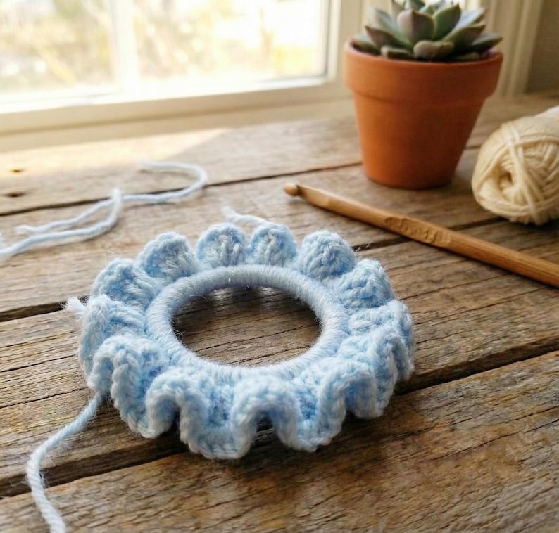 Op de afbeelding: Een lichtblauwe, handgemaakte gehaakte haarband met een gerimpelde rand. Het haaraccessoire is cirkelvormig, met een zichtbare elastische band in het midden. De achtergrond omvat een houten haaknaald, een bol wol en een vetplant in een pot.