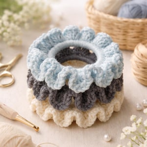 Op de afbeelding: Een stapel van drie handgemaakte gehaakte scrunchies in lichtblauw, grijs en cr&egrave;me. De scrunchies hebben een gerimpelde rand en zijn gemaakt van zacht garen. Een haaknaald, garen en schaar staan op de achtergrond.