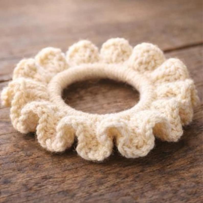 Op de afbeelding: Een cr&egrave;mekleurige, gehaakte haarband met een gefranjerd, bloemachtig ontwerp. Het haaraccessoire is cirkelvormig met een centrale opening en een getextureerde, handgemaakte uitstraling. De achtergrond is een houten oppervlak.