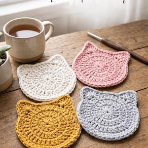 May include: Four handmade crochet cat coasters in white, pink, gold, and gray, arranged on a wooden surface. The coasters are cat-shaped with a circular design. The text "Crochet Cat Coaster" is at the top of the image.