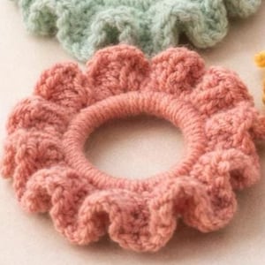 Op de afbeelding: Close-up van een roze gehaakte scrunchie met een gerimpelde rand. De scrunchie is cirkelvormig met een centrale opening en is gemaakt van zacht, getextureerd garen. Een lichtgroene scrunchie is zichtbaar op de achtergrond.