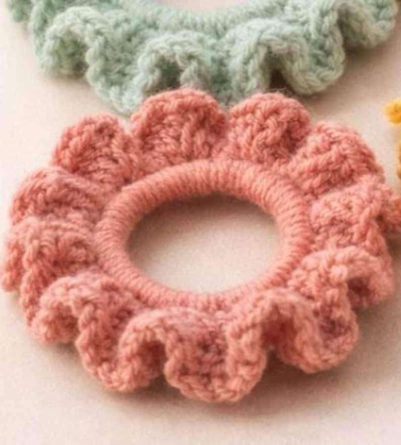 Op de afbeelding: Close-up van een roze gehaakte scrunchie met een gerimpelde rand. De scrunchie is cirkelvormig met een centrale opening en is gemaakt van zacht, getextureerd garen. Een lichtgroene scrunchie is zichtbaar op de achtergrond.