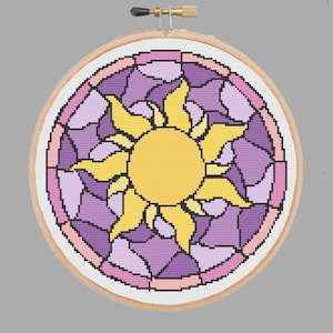 May include: A cross-stitch embroidery hoop featuring a stained glass design of a sun with yellow rays and a purple and pink background.