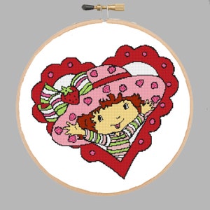 Puede incluir: Un marco rojo en forma de corazón con lunares blancos rodea una imagen de dibujos animados de Strawberry Shortcake. Lleva un sombrero rosa y verde con una fresa en la parte superior y una camisa a rayas rosa y verde.