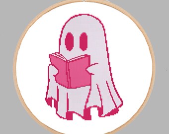 May include: A cross-stitch embroidery project in a circular wooden hoop featuring a pixel art design of a white ghost with dark pink eyes holding an open dark pink book. The ghost has a simple, rounded shape and is centered on white fabric.