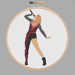 May include: Cross-stitch pattern of a woman in a black and red outfit singing into a microphone. The pattern is in a circular embroidery hoop.