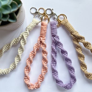 May include: Four handmade macrame keychains in cream, peach, lavender, and beige. Each keychain features a twisted rope design and a gold key ring.