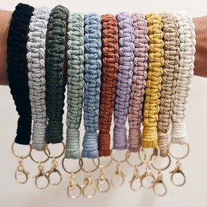 May include: A set of 11 colourful macrame keychains with gold key rings. The keychains are made of different colours of yarn, including black, white, green, blue, brown, purple, yellow, and beige.