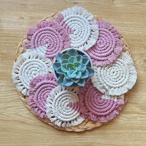 May include: A set of handmade macramé coasters in pink and cream, surrounding a small succulent in a ceramic pot. The coasters feature a spiral design with fringed edges, placed on a woven placemat, creating a bohemian style.