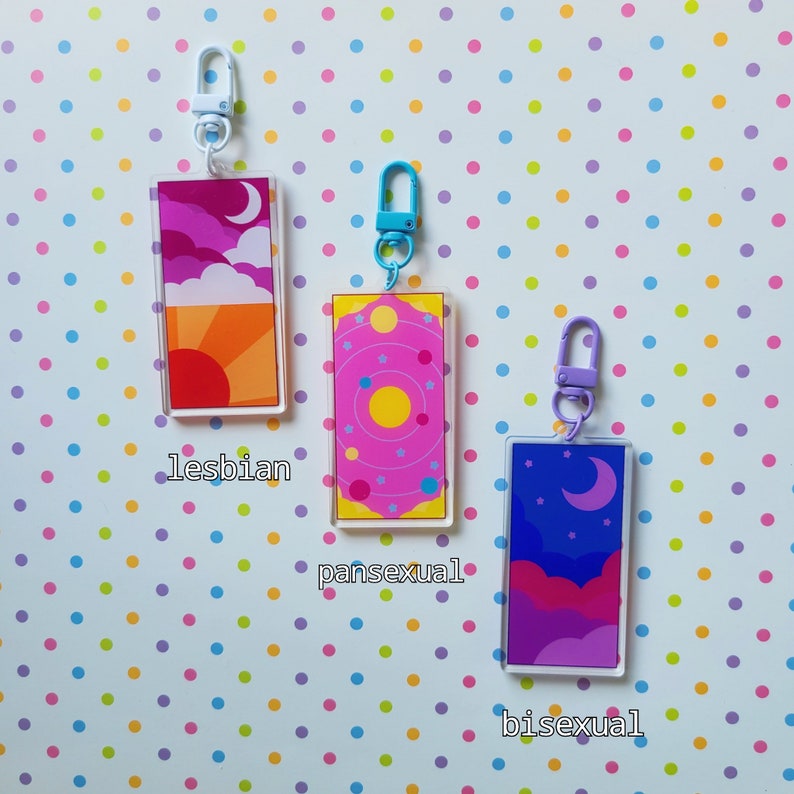 May include: Three colorful acrylic keychains with different designs representing different sexual orientations. The keychain on the left has an orange, yellow, and pink sunset design with a crescent moon and the word "lesbian" below it. The middle keychain has a pink background with a yellow sun and a circle of planets and the word "pansexual" below it. The keychain on the right has a purple, pink, and blue night sky design with a crescent moon and the word "bisexual" below it.