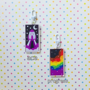 May include: Two colorful keychains with white metal rings. The keychain on the left features a purple rocket ship with white flames and stars against a dark blue background. The keychain on the right features a rainbow with white clouds and stars against a dark blue background. The text "ace" is below the left keychain and the text "progress" is below the right keychain.
