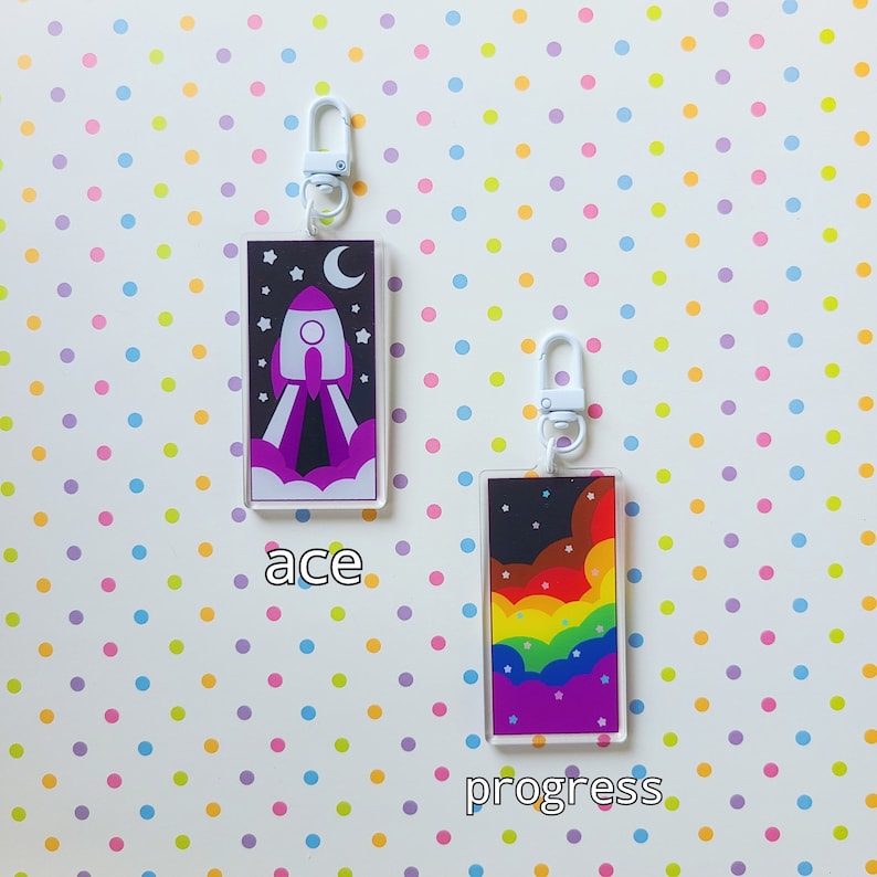 May include: Two colorful keychains with white metal rings. The keychain on the left features a purple rocket ship with white flames and stars against a dark blue background. The keychain on the right features a rainbow with white clouds and stars against a dark blue background. The text "ace" is below the left keychain and the text "progress" is below the right keychain.