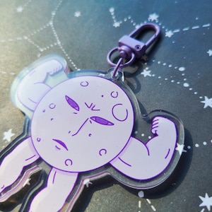 May include: Purple and white acrylic keychain featuring a cartoon moon with a muscular body flexing its biceps. The moon has a serious expression with purple eyes and a small mouth.