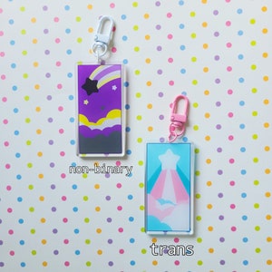 May include: Two colorful acrylic keychains with a star and clouds design. The keychain on the left is purple with a rainbow stripe and the text "non-binary". The keychain on the right is blue with a pink and white stripe and the text "trans".