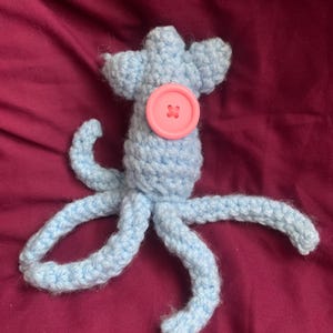 May include: A light blue crocheted octopus toy with eight tentacles and a pink button. The handmade plushie is on a maroon background. The octopus has a round body and a head with two small protrusions.